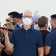 Apple CEO Tim Cook stands in the middle of a throng of people wearing masks, holding a laptop in one hand and staring at the camera.
